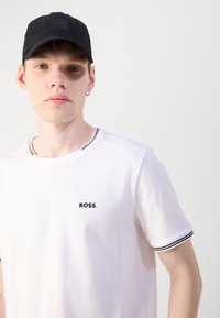 Balta crew tipo marškinėliai su juodu "BOSS" logotipu, su juodai balto dryžuoto apdailos ant rankovių. Juoda kepurė su logotipo detale.