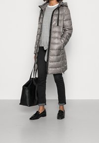 Osoba ubrana w szary puchowy płaszcz, szary prążkowany top, czarne materiałowe spodnie podwinięte na dole, czarne mokasyny, trzymająca dużą czarną torbę na ramię, stojąca na szarej podłodze.