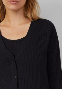 Zwarte geribde cardigan met een V-hals en knoopsluiting, gelaagd over een bijpassende geribde tanktop, met een gladde textuur.