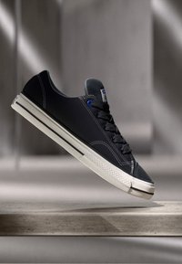 Black canvas sneaker with white rubber sole, featuring blue accents, six eyelets, and contrasting stitching. Low-cut design for casual wear.