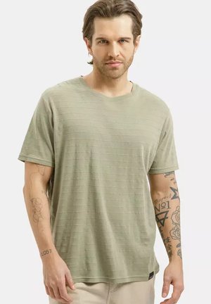 Mężczyzna o średniej długości włosach, ubrany w jasnozieloną koszulę w paski z krótkim rękawem, z widocznymi tatuażami na obu ramionach.