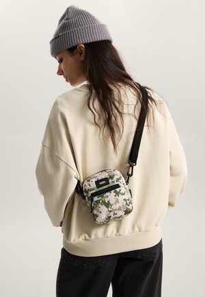 Bolso mini de camuflaje con dos cremalleras, herrajes negros y correa ajustable, llevado cruzado sobre un suéter de gran tamaño de color claro.