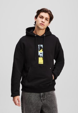 Schwarzer Kapuzenpullover mit einer Fronttasche, ausgestattet mit einem farbenfrohen Grafikdruck und gelben Akzenten. Glatte Textur, Kordeln an der Kapuze.