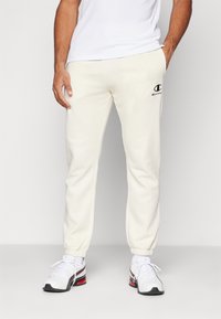 Pantalons joggers crème en tissu doux, dotés d'une taille élastique et de poignets élastiques, avec un logo brodé noir sur le devant gauche.