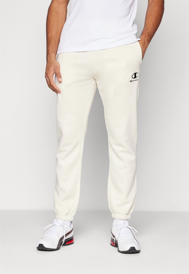 Pantalons joggers crème en tissu doux, dotés d'une taille élastique et de poignets élastiques, avec un logo brodé noir sur le devant gauche.