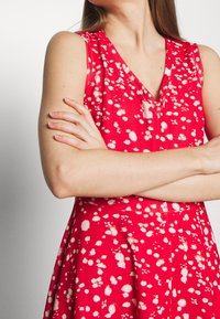 Mujer con los brazos cruzados, vistiendo un vestido sin mangas de color rojo con patrones blancos dispersos, mostrando desde el cuello hasta la cintura.