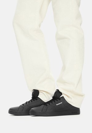 Čierne kožené tenisky majú nízky strih, okrúlu špičku, šnurovanie a jemné logo na boku. Nosia sa s krémovými nohavicami.