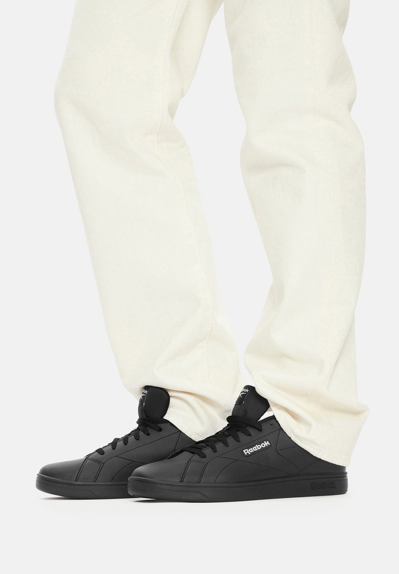 Čierne kožené tenisky majú nízky strih, okrúlu špičku, šnurovanie a jemné logo na boku. Nosia sa s krémovými nohavicami.