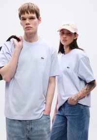 Ανοιχτόχρωμα λιλά βαμβακερά t-shirts με μικρό πράσινο λογότυπο, συνδυασμένα με μπλε τζιν. Ένα μοντέλο φοράει ροζ καπέλο με λογότυπο σε γραφή.