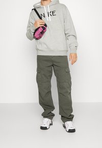 Šedá mikina s kapucí a černým logem "NIKE", olivově zelené kalhoty s kapsami a bílé boty s červenými detaily. Růžová taška přes rameno.