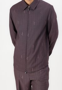 Zip-up jacket in dark purple fabric with vertical stripes. Features a pointed collar, subtle branding, and side pockets.