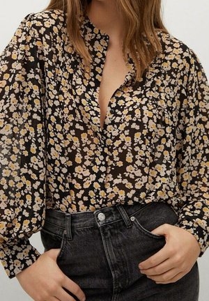 Femme portant un chemisier noir à fleurs blanches et jaunes et un jean noir taille haute, les mains reposant près des poches.