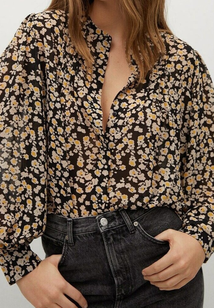 Femme portant un chemisier noir à fleurs blanches et jaunes et un jean noir taille haute, les mains reposant près des poches.