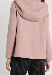 Ljusrosa hoodie-jacka i en texturerad tyg med en stor huva, i kombination med svarta byxor med smala ränder, som ger en avslappnad passform.