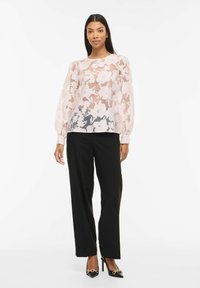 Femme portant un chemisier floral transparent à manches longues, un pantalon noir à jambes larges et des talons pointus noirs avec un détail chaîne en or, debout sur un fond blanc.