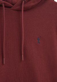 Felpa color mogano con una superficie texturizzata, caratterizzata da una piccola figura blu ricamata che tiene una mazza da golf e cordini abbinati.