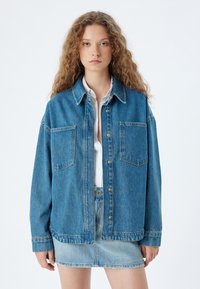 Blå denimjacka med avslappnad passform, som har två bröstfickor, knäppning med knappar och krage. Bärs över en vit skjorta och en denimkjol.