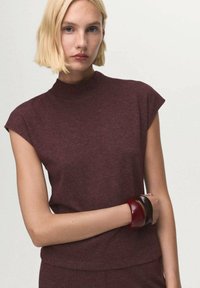 Burgundy, short-sleeved top with a high neck made from a soft fabric. Features a fitted silhouette. Three large bracelets in varying colors.