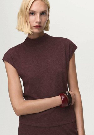 Top bourgogne à manches courtes avec col montant, fabriqué dans un tissu doux. Présente une silhouette ajustée. Trois grands bracelets de couleurs variées.