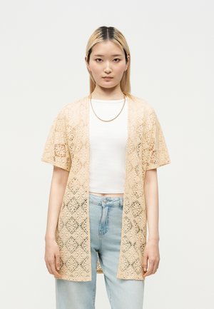 Ung kvinde med lige blondt hår bærer en beige blondecardigan over en hvid cropped top og lyseblå jeans, stående mod en ensfarvet baggrund.