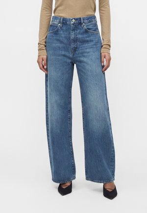 Personne portant un jean bleu taille haute à jambes larges, un haut beige à manches longues et des chaussures noires à bout pointu sur fond blanc.