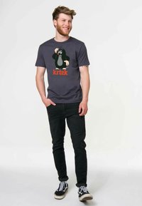 Young man in dark jeans and sneakers wearing a gray T-shirt with a cartoon mole and the word "krtek" smiling and looking right.