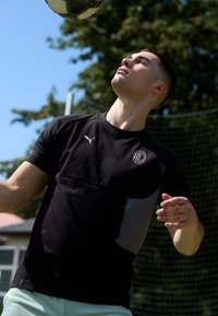 Jersey deportivo de manga corta negra con panel gris, con un logo en el pecho y el hombro. El fondo muestra vegetación y una red de portería.