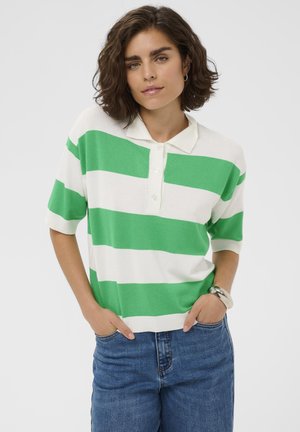 Ženska s kratkimi kodrastimi lasmi, oblečena v zeleno-belo črtasto polo majico in modre kavbojke, stoji z rokami v žepih ob belem ozadju.