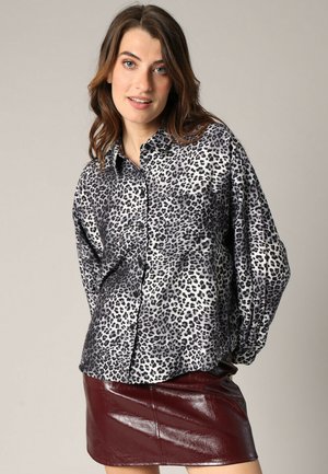 Jeune femme portant un blouson à manches longues en imprimé léopard et une mini-jupe en cuir rouge foncé brillant, se tenant devant un fond uni.