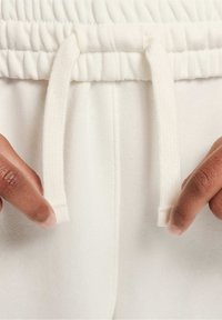 Bílé tepláky mají elastický pas se šňůrkami na stažení. Tkanina vypadá měkce a na detailním záběru je vidět ruka, jak drží tkaninu.