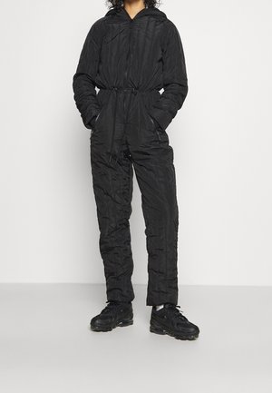 Osoba ubrana w czarny pikowany kombinezon z kapturem, zamkiem z przodu, regulowanym pasem na sznurek, kieszeniami i czarnymi trampkami na jednolitym tle.