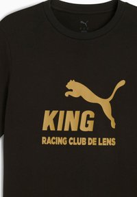 Sort T-shirt med gyldent tekst: "KING" over "RACING CLUB DE LENS" med et springende Puma-logo. Fokuseret på sport og holdånd.