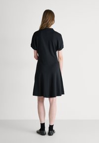 Robe noire à manches courtes avec une silhouette évasée, tissu lisse, texture subtile et col. Portée avec des chaussettes foncées et des chaussures plates.