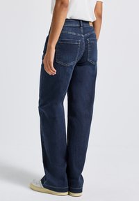 Jean en denim bleu foncé avec une coupe droite, deux poches arrière et une texture subtile. Porté avec des baskets blanches texturées.