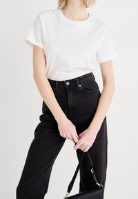 Vit bomull t-shirt med korta ärmar, rund halsringning, ihop med högmidjade svarta denimjeans. Personen håller en liten svart handväska.