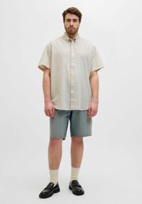 Chemise à manches courtes, boutonnée en tissu beige clair. Portée avec un short en denim bleu clair et des mocassins noirs, complétée par des chaussettes couleur crème.