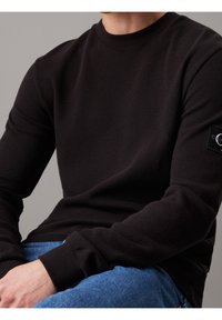 Black long-sleeve top with ribbed texture, crew neckline, and a logo patch on the left sleeve. Paired with blue denim jeans.