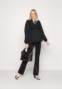 Blusa preta com babados, combinada com calças pretas brilhantes e sandálias de salto alto. Os acessórios incluem uma pequena mala de ombro preta texturizada.