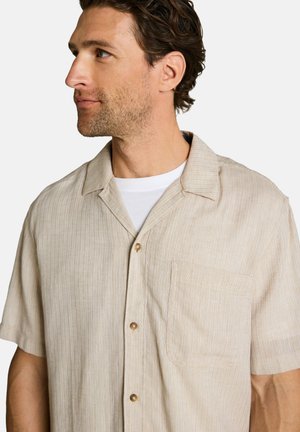 Man with short brown hair wearing a beige, short-sleeve, button-up shirt over a white t-shirt, looking to the left.