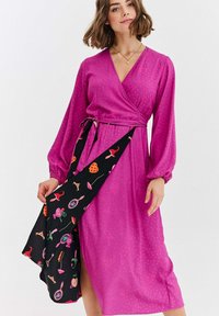 Różowa sukienka kopertowa z długimi balonowymi rękawami, ozdobiona czarnym wzorzystym szalikiem z kolorowymi motywami. Gładka faktura i długość do kolan.