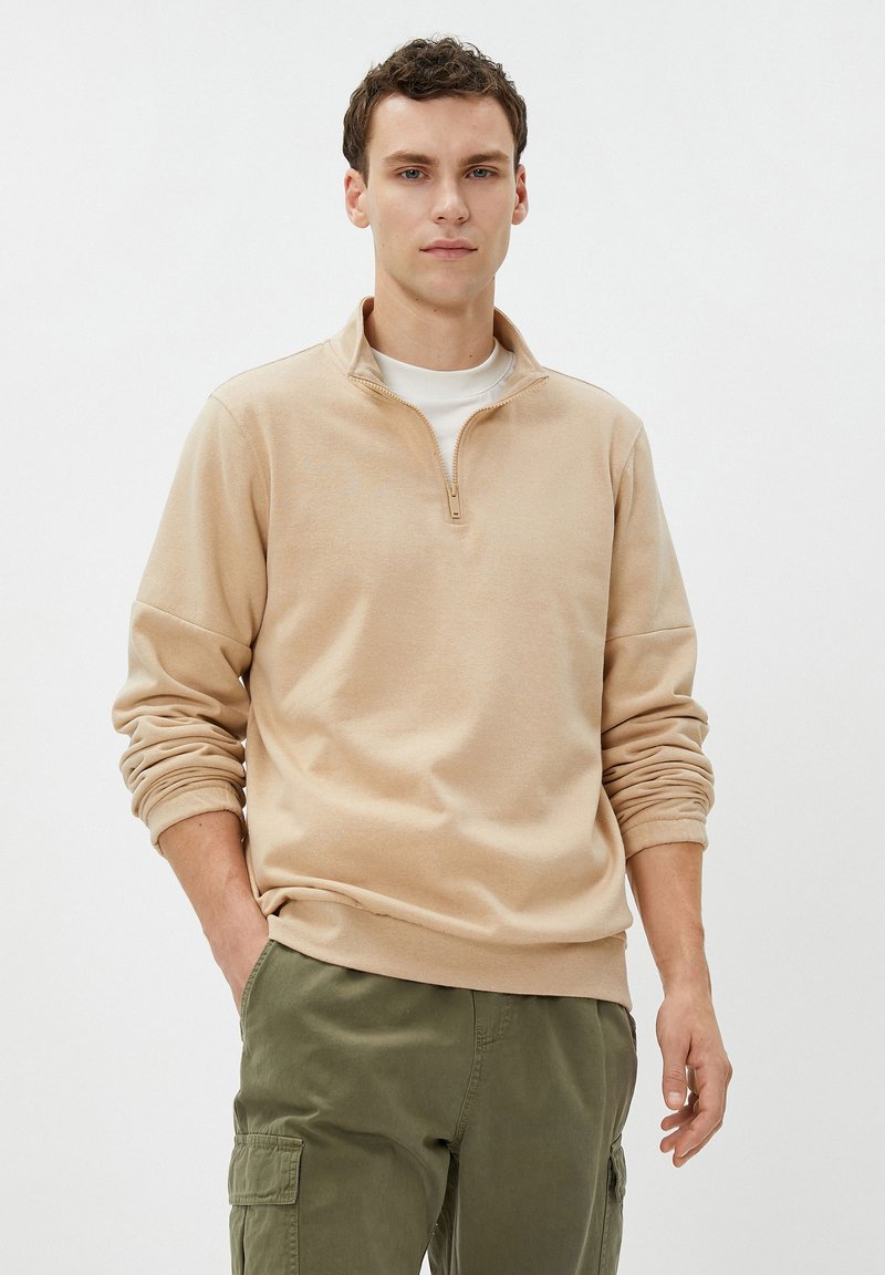 Sudadera beige con cremallera de un cuarto y mangas largas, con puños acanalados y una textura suave, llevada sobre una camiseta blanca, combinada con pantalones verde olivo.