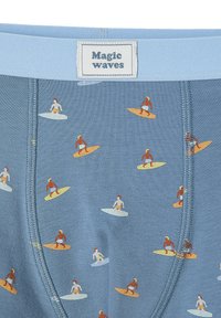 Blauer Stoff mit einem Muster aus kleinen Surfern auf gelben und orangefarbenen Surfboards, mit einem "Magic Waves" Etikett am Bund.