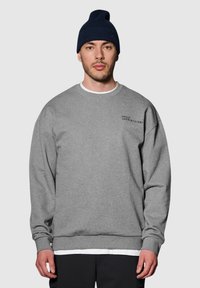 Grijze sweatshirt met ronde hals, geribbelde boorden en onderrand. Bevat kleine zwarte tekst op de bovenste linker borst. Gekleed met een marineblauwe beanie.
