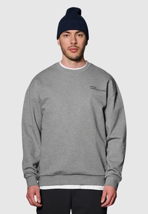 Grauer Pullover mit Rundhalsausschnitt, gerippten Bündchen und Saum. Enthält kleinen schwarzen Text auf der oberen linken Brust. Getragen mit einer marineblauen Mütze.
