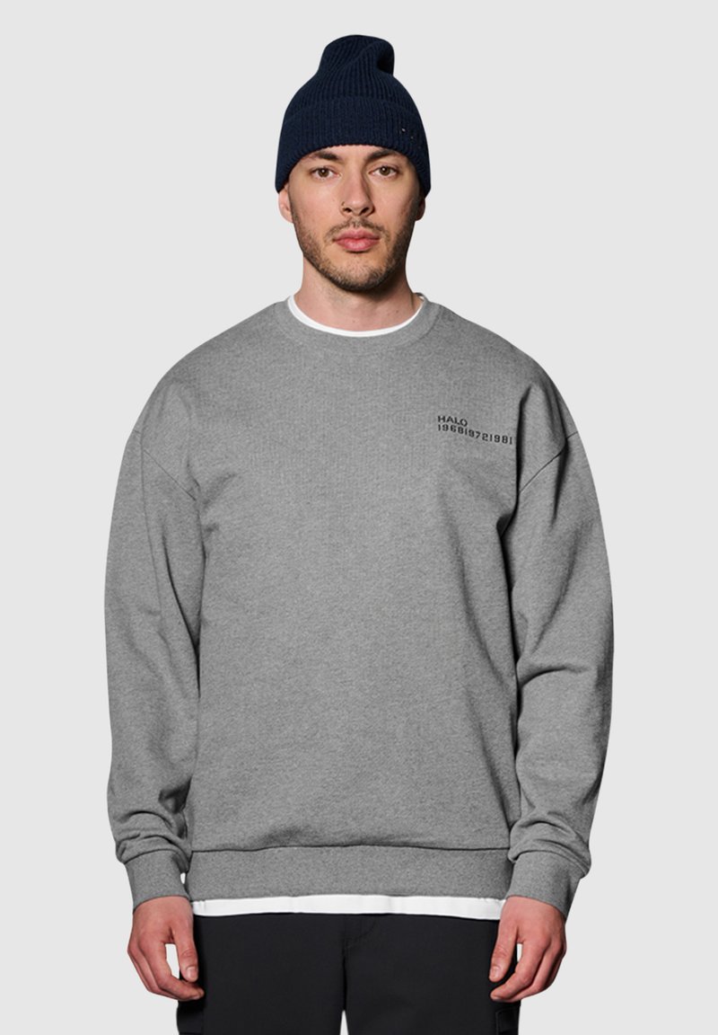 Grijze sweatshirt met ronde hals, geribbelde boorden en onderrand. Bevat kleine zwarte tekst op de bovenste linker borst. Gekleed met een marineblauwe beanie.