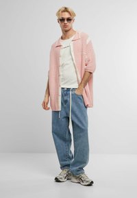 Overdimensjonert rosa og hvit rutete skjorte med korte ermer, hvit T-skjorte under, avslappede blå jeans og grå joggesko med svarte detaljer.
