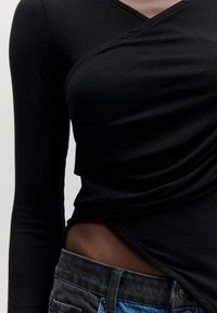 Schwarzes, geripptes Langarmoberteil mit V-Ausschnitt, das eine drapierte Textur an der Taille aufweist, kombiniert mit dunklen Jeans.