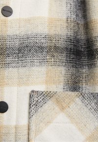 Primer plano de una tela de cuadro en tonos beige, negro y blanco, con un bolsillo frontal y botones negros mate etiquetados como Bershka.