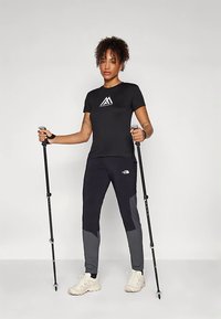 Svart kortermet skjorte med hvit logo, kombinert med tettsittende svarte og grå bukser. Bruker turgåerstaver; fottøyet er lyse farger og sporty.