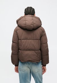 Persoon met gevlochten haar, gekleed in een bruin gewatteerd jack met een grote capuchon en lichtblauwe spijkerbroek, met de rug naar de camera.